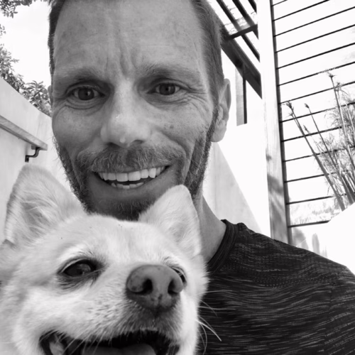 a black and white photo of a man with short hair and facial hair, with a smiling light-colored dog in the foreground